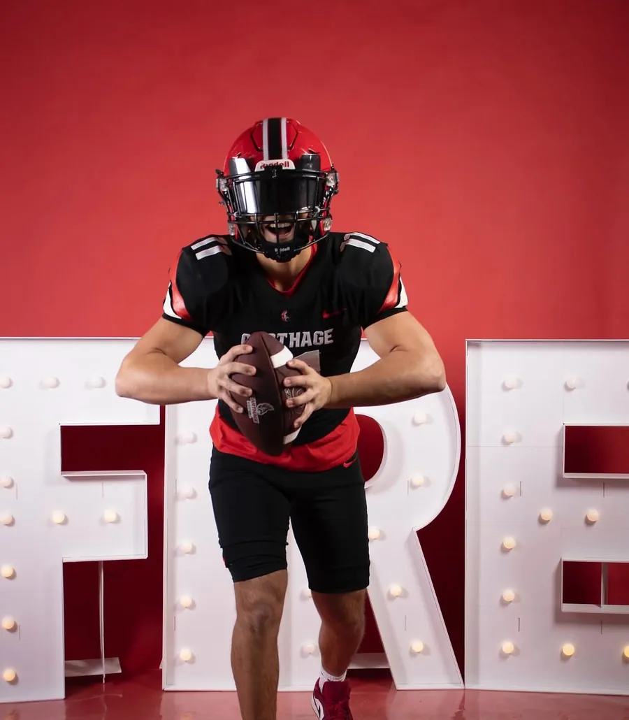 Will in a black Carthage jersey holding a football in front of lighted marquee letters
